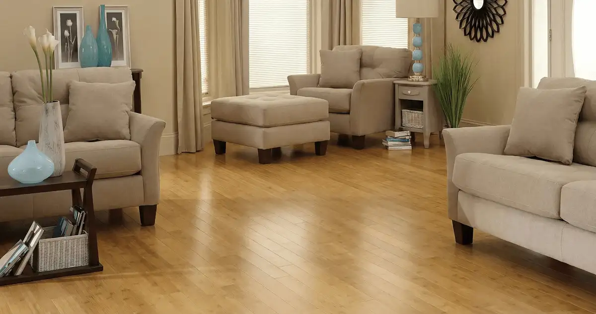 A living room with professionally installed bamboo flooring, beige sofas, a matching ottoman, a side table with a blue lamp, vases, framed pictures, and large windows with blinds and curtains in Tampa, FL